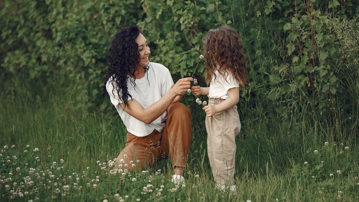 A mother and daughter bond outdoors in a lush green field during summer, sharing flowers.