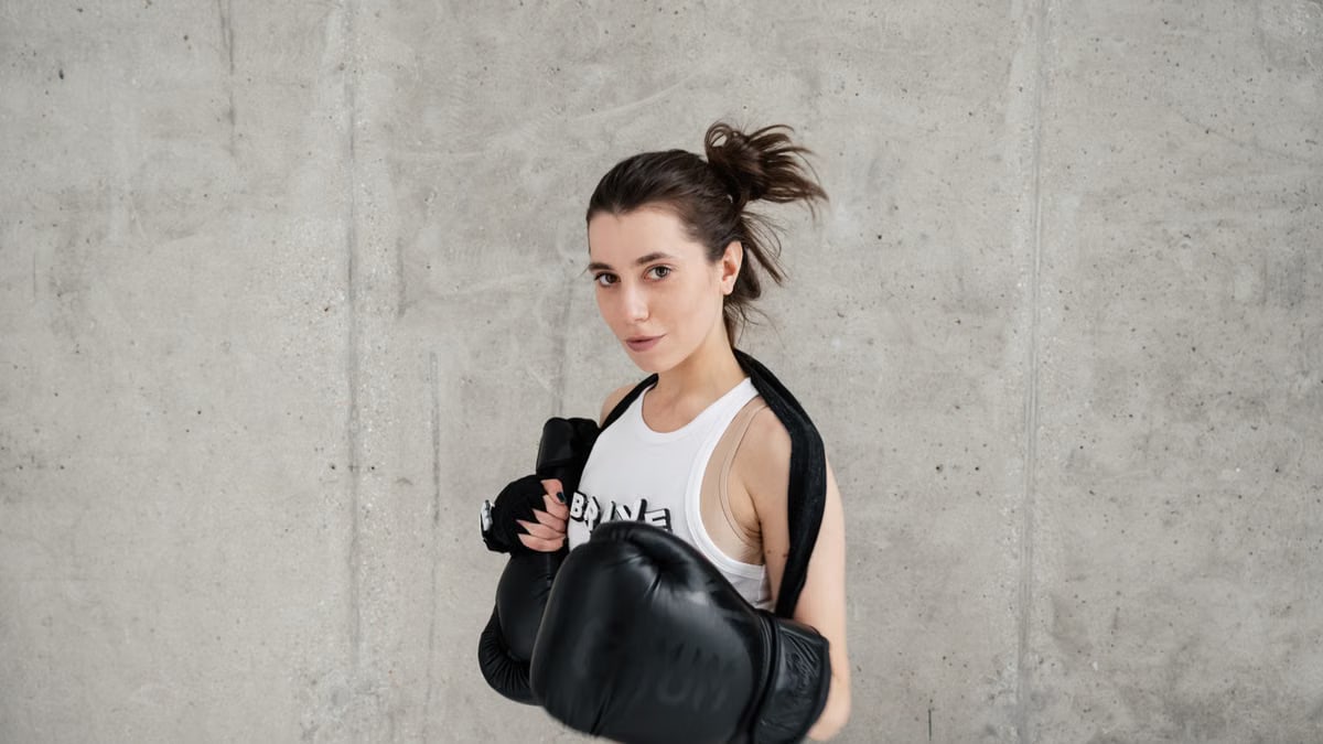 Confident young woman posing with boxing gloves against a concrete wall, showcasing style and sportiness.